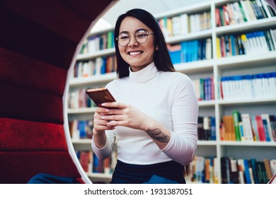 Content Asian woman in glasses surfing mobile phone in tattooed hand sitting in round soft bench in wall of library and looking at camera with smile