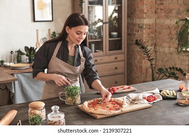 Content attractive young woman in apron standing at counter and preparing pizza with herbs in kitchen - Powered by Shutterstock