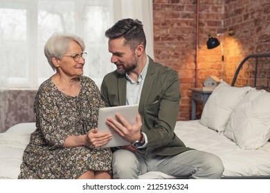 Content Caucasian Man Showing New Helpful Application On Tablet Computer To His Elderly Mother. High Quality Photo