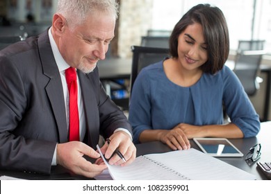 Content Coach Reviewing Notes Of Female Company Newcomer. Middle Aged Man In Tie And Young Indian Woman In Casual Wear Thumbing Notebook In Coworking Space. Business Coaching And Mentorship Concept