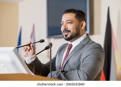 Content confident Middle Eastern politician in gray suit standing at tribune podium and speaking into microphone while presenting report at conference - Shutterstock ID 2065631840