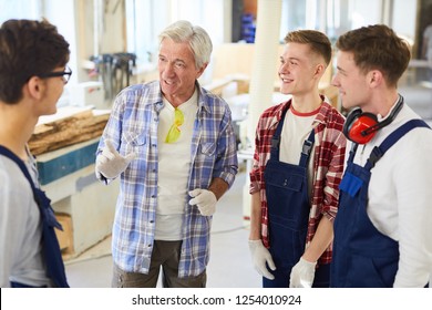 Content Confident Skilled Carpenter In Work Gloves Standing In Circle Of Young Interns And Gesturing Hands While Sharing Experience With Young Workers In Modern Workshop