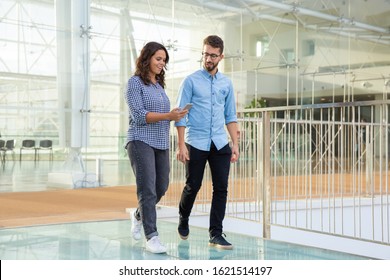 Content Couple Walking And Using Smartphone. Cheerful Young Man And Woman Walking Inside Office Building And Using Mobile Phone. Technology Concept