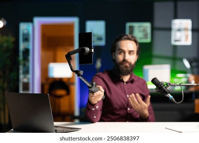Content creator sitting at table talking in front on smartphone attached to selfie stick in home studio interior, chatting with fans. Man using phone camera to record vlog in neon lit apartment - Powered by Shutterstock - Get 15% off with code: PIKWIZARD15