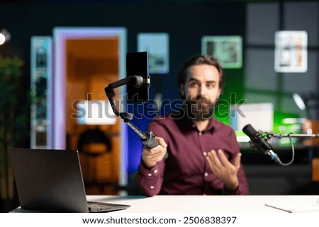 Content creator sitting at table talking in front on smartphone attached to selfie stick in home studio interior, chatting with fans. Man using phone camera to record vlog in neon lit apartment