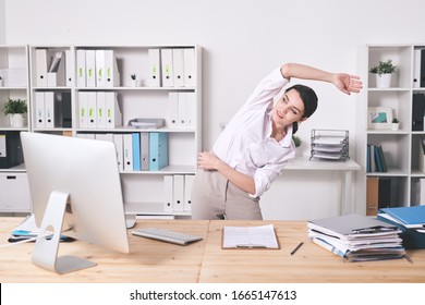 Content energetic young woman in blouse bending side and stretching arm while doing warm-up exercise in office