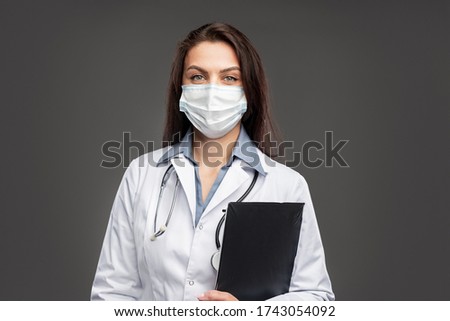 Content female physician wearing medical gown and protective mask standing on gray background with clipboard during COVID 19 pandemic and looking at camera