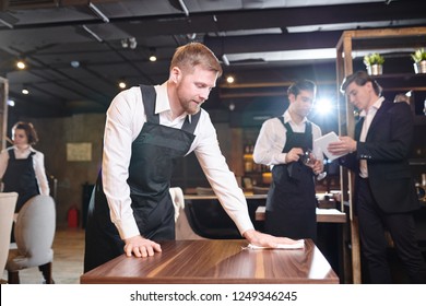 Content Handsome Young Bearded Waiter  In Black Apron Wiping Table With Cloth In Restaurant While Manager Showing Seating Chart To Waiter In Background