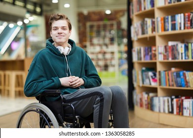 Content handsome young disabled student with headphones on neck siting in wheelchair and looking at camera in modern library or bookstore - Shutterstock ID 1363058285