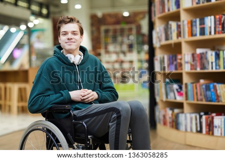 Content handsome young disabled student with headphones on neck siting in wheelchair and looking at camera in modern library or bookstore