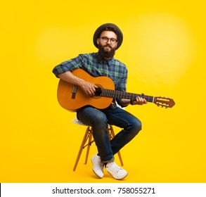 Content hipster man in hat and eyeglasses playing guitar sitting on chair on orange studio background. 