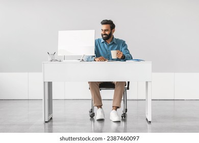 Content indian male professional with beard enjoying his coffee while working on computer at clean and minimalistic white office desk, full length - Powered by Shutterstock