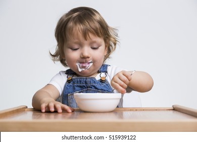 Content Little Guy Enjoying His Porridge Or Yogurt. He Is Wearing A White T-shirt And Blue Denim Overalls. 