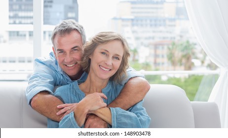 Content Man Hugging His Wife On The Couch Smiling At Camera At Home In The Living Room