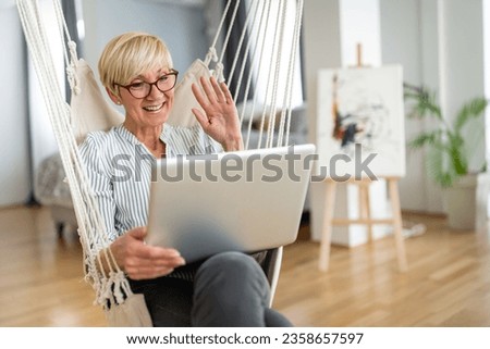 Content merry older Caucasian woman looking at laptop screen and waving during video call while sitting in rope swing at home.