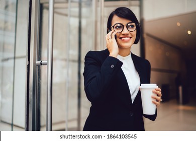 Moderne Geschäftsfrau in formeller Kleidung und Brille mit einer Tasse Kaffee aus dem Ausland im Café stehen und über Smartphone sprechen