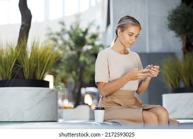 Content Pensive Young Woman With Tattoo On Arm Sitting On Bench In Shopping Center And Checking Messenger On Phone