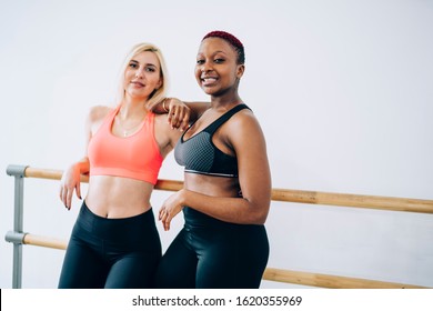 Content relaxed diverse sportswomen in activewear standing in white fitness center room and leaning on ballet barre while looking at camera