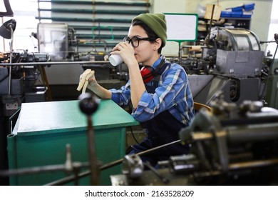 Content Relaxed Young Woman In Workwear Drinking Takeout Coffee And Eating Sandwich At Factory Workplace