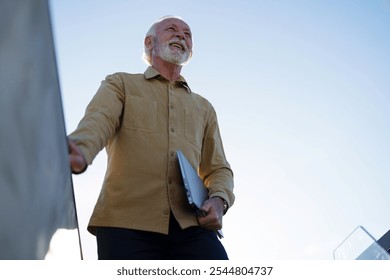 A content senior Caucasian male executive uses a laptop in a sunny outdoor setting, dressed in professional casual attire, embodying mobile work flexibility. - Powered by Shutterstock