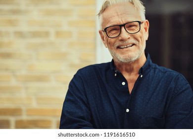 Content Senior Man With A Beard And Wearing Glasses Laughing While Standing Outside In Front Of His Home