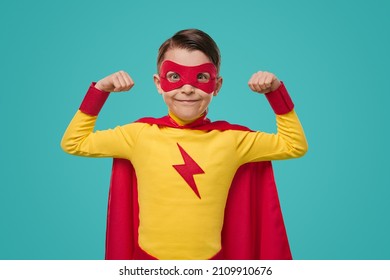 Content Superhero Kid In Red And Yellow Cape And Mask Demonstrating Power While Looking At Camera Against Blue Background In Studio