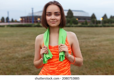 Content Sweaty Fit Female Runner With Towel On Neck And In Orange Top Standing In Field After Active Training In Summer And Looking At Camera 