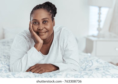 Content young African woman wearing pajamas lying on her bed with her hand on her chin while waking up in the morning - Powered by Shutterstock