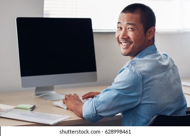 Content Young Asian Entrepreneur Looking Over His Shoulder While Working Alone On A Computer In A Clean, Modern Office