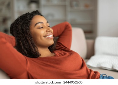 Content young black woman with a beaming smile reclines on a couch, her hands behind her head, eyes closed in serene repose, african american female resting at home - Powered by Shutterstock