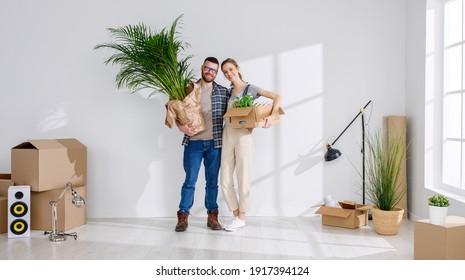 Content Young Family Couple Standing With Carton Boxes And Flowerpot In New Flat And Enjoying Relocation While Looking At Camera