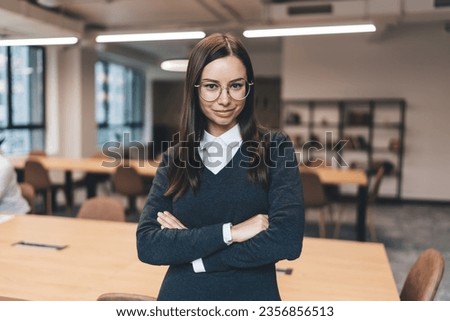 Content young female entrepreneur in elegant outfit and eyeglasses looking at camera and smiling while standing with folded arms in contemporary office