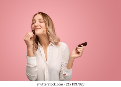 Content young female with long blond hair in white shirt smiling while enjoying smell of delicious chocolate with closed eyes against pink background