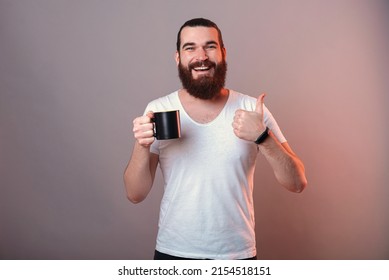 Content Young Man Is Holding A Black Mug While Showing Thumb Up For Like. Studio Shot Over Grey Background With Red Light.