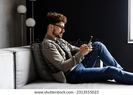 Content young man relax on sofa use smartphone spend free time online in social network chat at web app play easy game online. Smiling guy hold modern cell dial number on screen make call to friend
