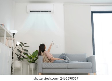 Content young woman lounges on a sofa, holding a remote to adjust her wall-mounted air conditioner, appreciating the cool air in a modern, bright living room setting - Powered by Shutterstock