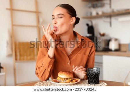 Contented caucasian lady licking her fingers while eating delicious french fries and hamburger, enjoying junk food at home in kitchen interior, copy space