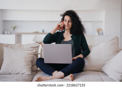 Contented, Curly-haired, Smiling Spanish Woman, Sitting On The Couch With A Laptop, Talking By  Phone, Looking Away, Received Pleasant News. Beautiful Curly Italian Woman Talking To Her Husband.