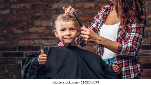 Contented cute preschooler boy shows thumbs up while getting a haircut. Children hairdresser with scissors and comb is cutting little boy in the room with loft interior.