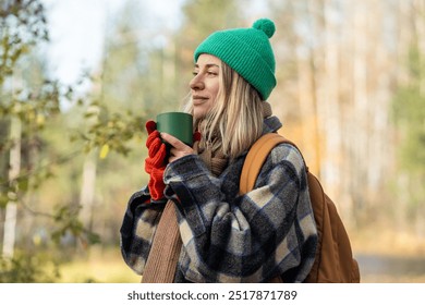 Contented, peaceful woman drinking beverage to keep warm while camping journey tourism through forest in sunny cool weather. Happy woman outside holding cup, inhaling delicious aroma with pleasure.