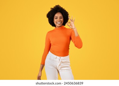 Contented woman with curly hair in orange turtleneck and white pants makes the OK sign, flashing a radiant smile, with a vibrant yellow backdrop highlighting her cheerful demeanor - Powered by Shutterstock