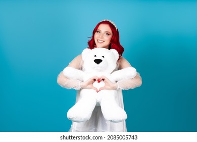 a contented woman in love in a white dress with red hair gently hugs a white teddy bear and makes a heart symbol from her fingers isolated on a blue background.