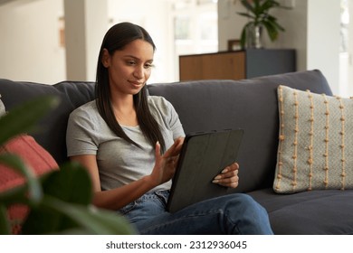Contented young biracial woman in casual clothing using digital tablet on sofa in living room