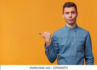 A Contentedly Pompous, Arrogant Guy Isolated Over Yellow Background Casually Draws Your Attention To What Is On The Left, Shows With Thumb Finger On The Empty Space To Copy.