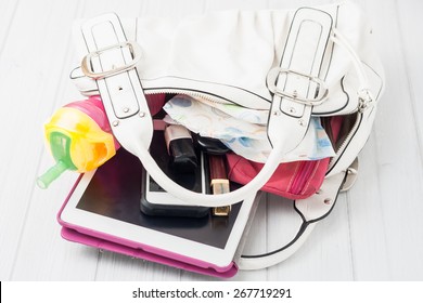 Contents Of A Woman's White Shoulder Bag On White Background. Contents Include: Tablet, Telephone, Lipstick, Nail Polish, Wallet, Baby Bottle And A Diaper.