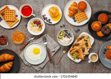 Continental Breakfast Captured From Above (top View, Flat Lay). Coffee, Tea, Croissants, Jam, Egg, Pancakes, Maffins And Oatmeal. Wooden Background. Family Breakfast Table.