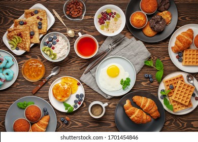 Continental Breakfast Captured From Above (top View, Flat Lay). Coffee, Tea, Croissants, Jam, Egg, Pancakes, Maffins And Oatmeal. Wooden Background. Family Breakfast Table.