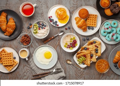 Continental Breakfast Captured From Above (top View, Flat Lay). Coffee, Tea, Croissants, Jam, Egg, Pancakes, Maffins And Oatmeal. Wooden Background. Family Breakfast Table.