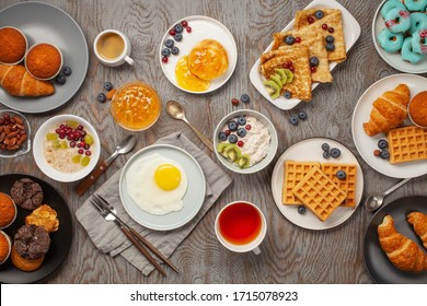 Continental Breakfast Captured From Above (top View, Flat Lay). Coffee, Tea, Croissants, Jam, Egg, Pancakes, Maffins And Oatmeal. Wooden Background. Family Breakfast Table.