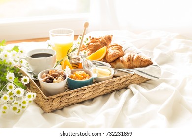 Continental breakfast on white bed sheets. Coffee, orange juice, croissants, jam, honey and flowers on wicker tray. Romantic countryside morning scenery, window as background.